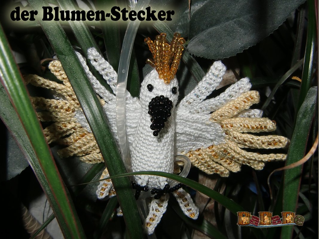 Gehäkelter weißer Kakadu-Blumenstecker mit goldfarbenen Perlen am Kamm und an den Flügeln zwischen grünen Pflanzenblättern.