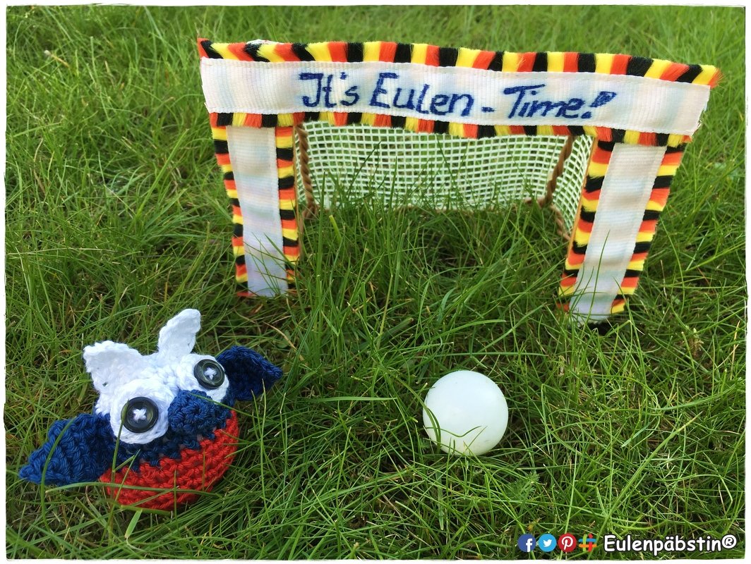 Häkeleule auf Gras neben Mini-Fußballtor mit schwarz-rot-gelben Bändern und weißem Ball