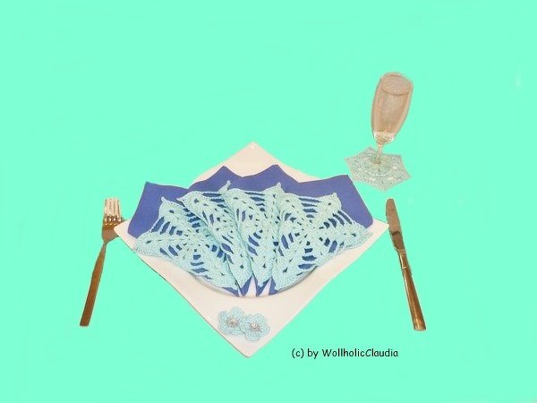 Gedeck mit hellblauer gehäkelter Serviettenauflage, Untersetzer und kleinen Blüten auf weißem Teller und blauer Serviette, Besteck und Glas.