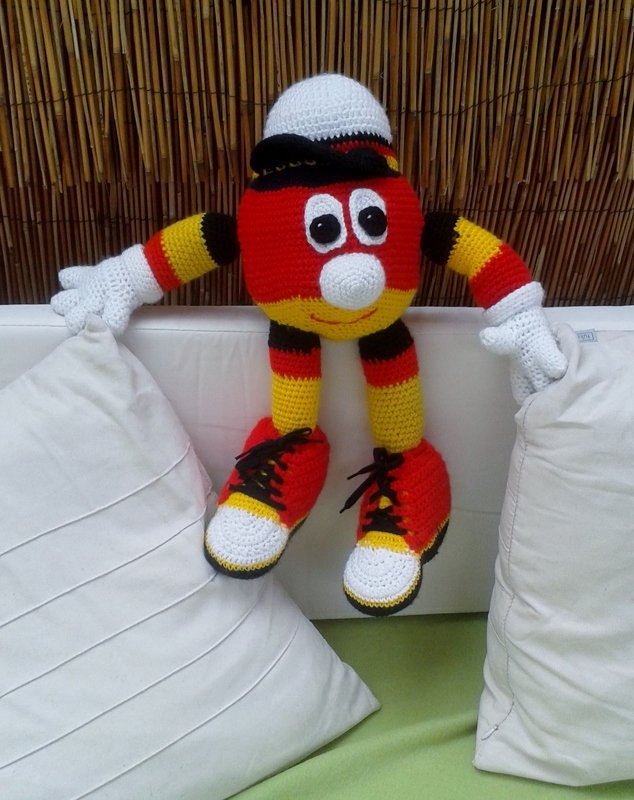 Gehäkelte Amigurumi-Figur in Schwarz-Rot-Gelb mit Mütze, Handschuhen und Turnschuhen auf weißem Sofa