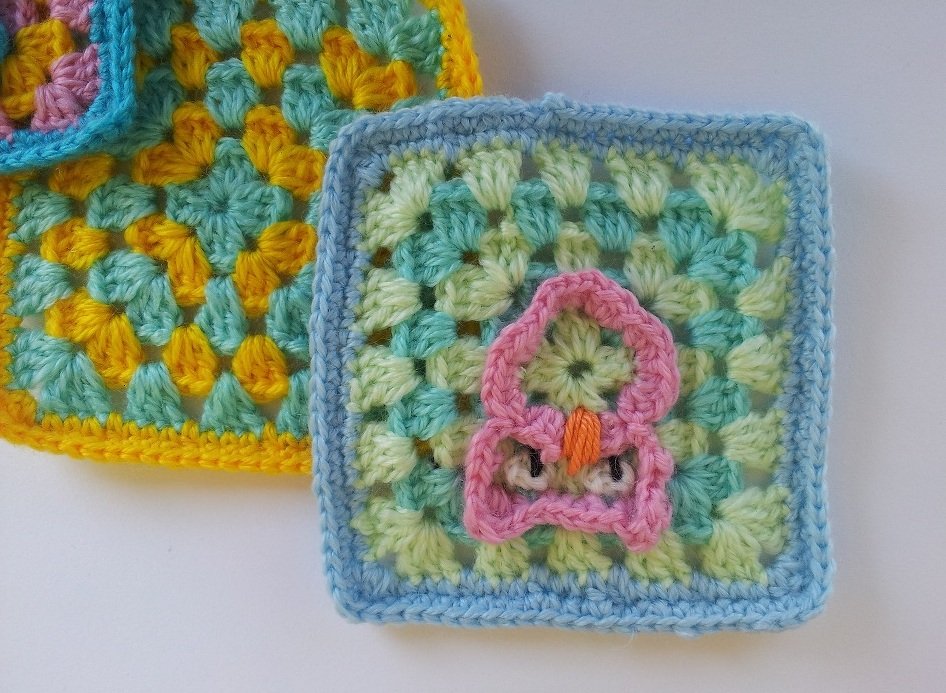 Handgehäkeltes Granny Square mit hellblauem Rand, mintgrün-hellgelbem Muster und einer kleinen rosa Eulen-Applikation mit orangem Schnabel; ein weiteres Granny Square ist teilweise sichtbar.