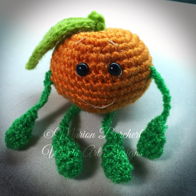 Gehäkelte orangefarbene Amigurumi Frucht mit schwarzen Augen, gesticktem Mund, grünem Blatt und grünen Beinchen