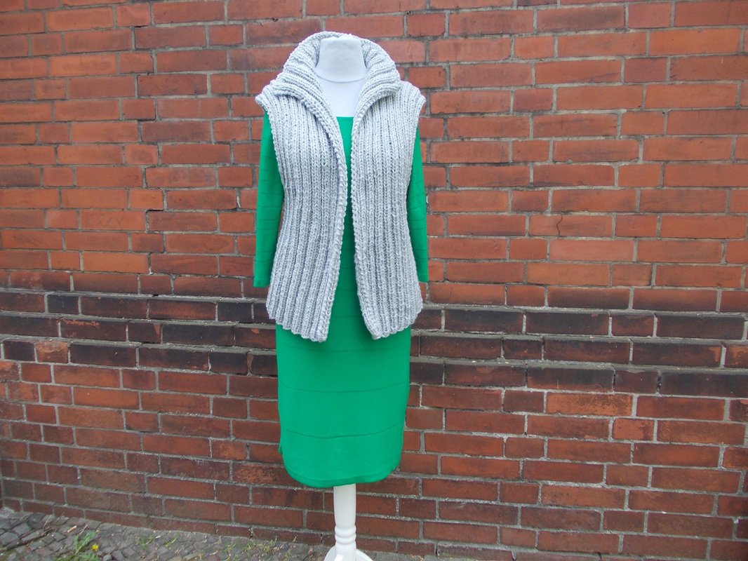 Mannequin mit grauer ärmelloser Strickweste mit Kapuze über grünem Kleid vor roter Backsteinwand