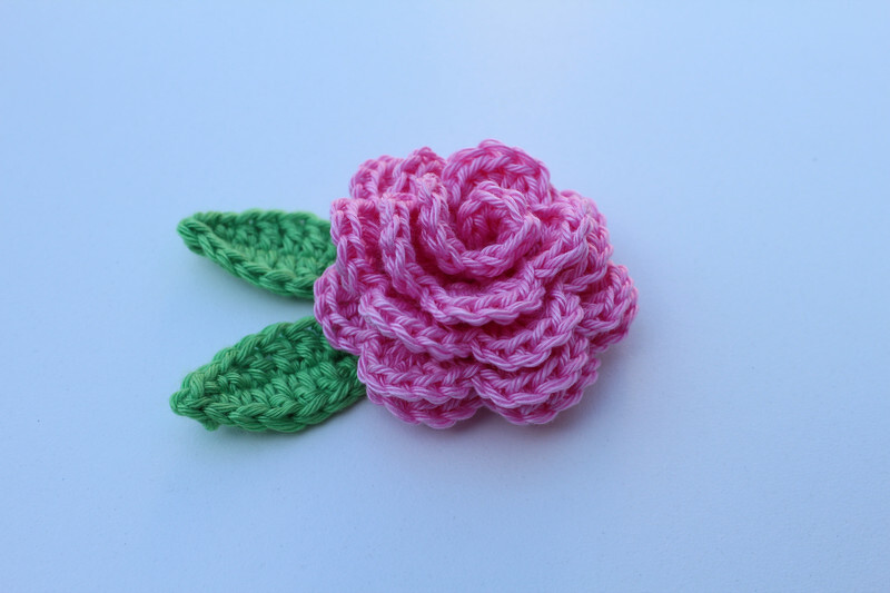 Gehäkelte pinke Rose mit zwei grünen Blättern auf hellem Hintergrund
