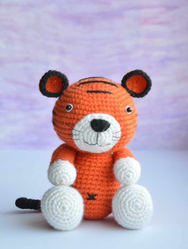 Front view of a crocheted orange amigurumi tiger showing stripes, white paws, and tail