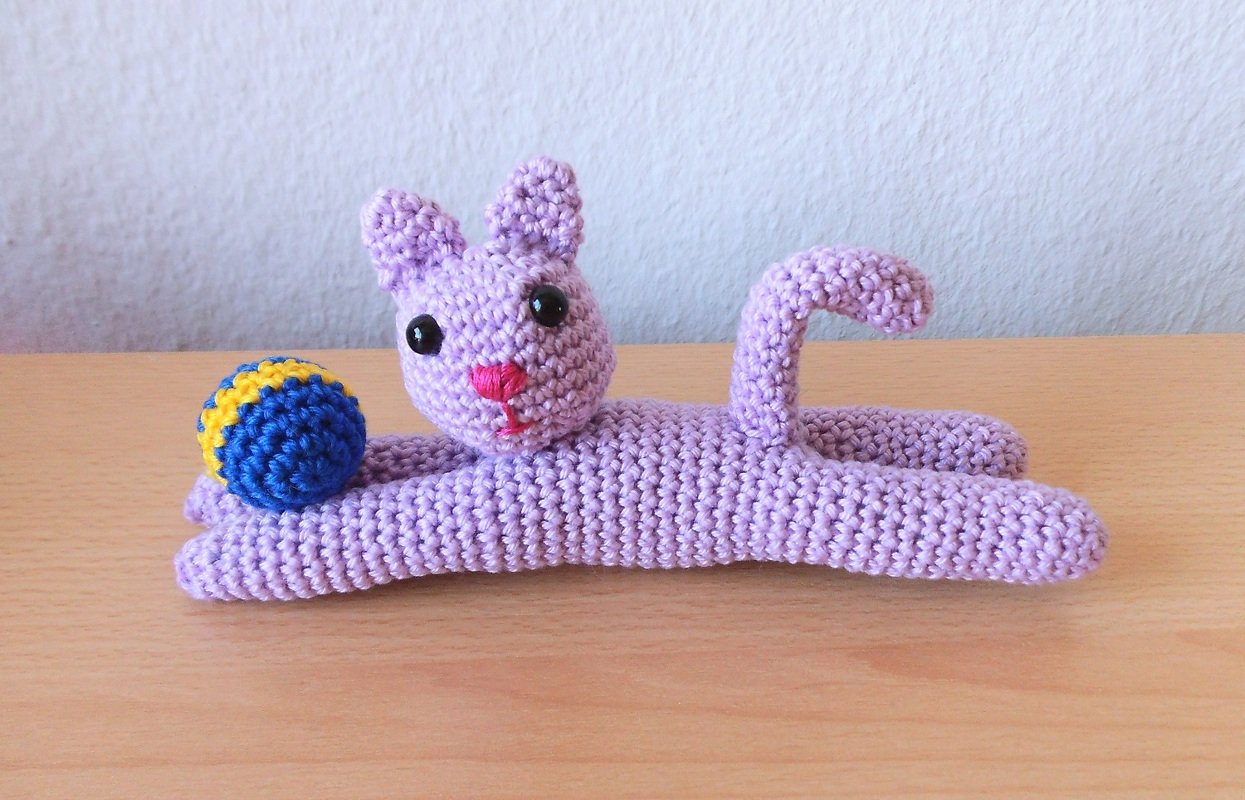 Lavendelfarbene gehäkelte Katze liegend auf Holztisch mit blau-gelbem Ball
