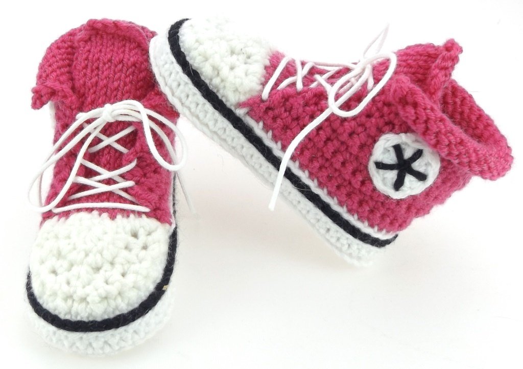 Nahaufnahme zweier gehäkelter Babyschuhe in Pink, Weiß und Schwarz mit Schnürsenkeln und rundem Seitenemblem
