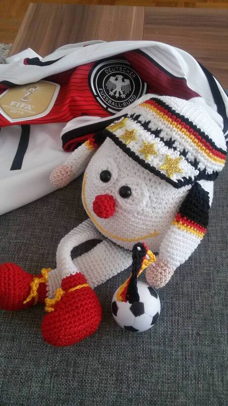 Weißes gehäkeltes Amigurumi mit schwarz-rot-goldener Mütze, goldenen Sternen, roten Schuhen und kleinem schwarz-weißem Fußball