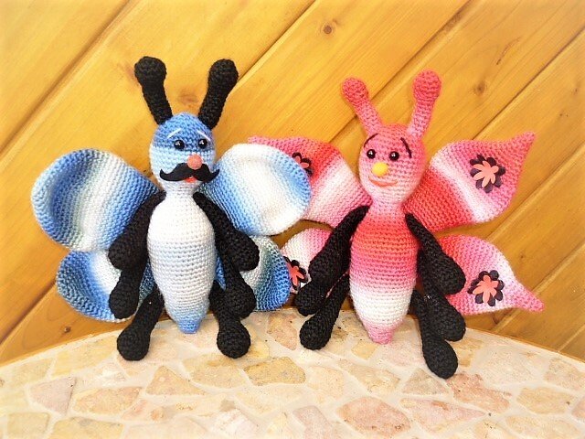 Zwei gehäkelte Amigurumi‑Schmetterlinge (blau‑weiß mit Schnurrbart, rosa‑weiß mit Blumen) vor Holzverkleidung.