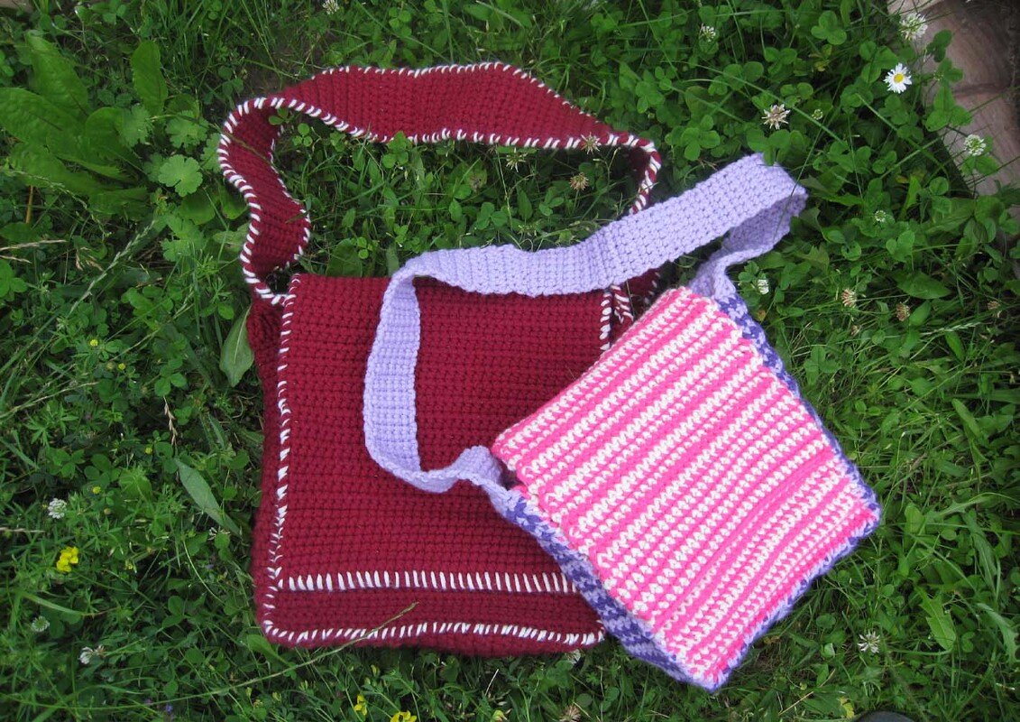 Zwei gehäkelte Umhängetaschen: burgunderrote Tasche mit weißer Kante und rosa-weiß gestreifte Tasche mit lila Träger auf Gras