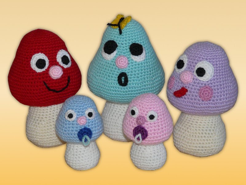 Fünf gehäkelte Amigurumi-Pilze (rot, türkis, violett, zwei kleine blau/rosa) mit weißen Stielen und Gesichtsausdrücken