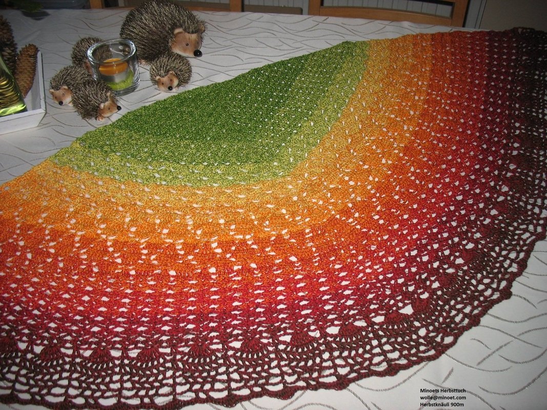 Halbrundes gehäkeltes Tuch mit Farbverlauf von Grün über Gelb und Orange bis Rotbraun auf einem Tisch, dekoriert mit Igel‑Figuren