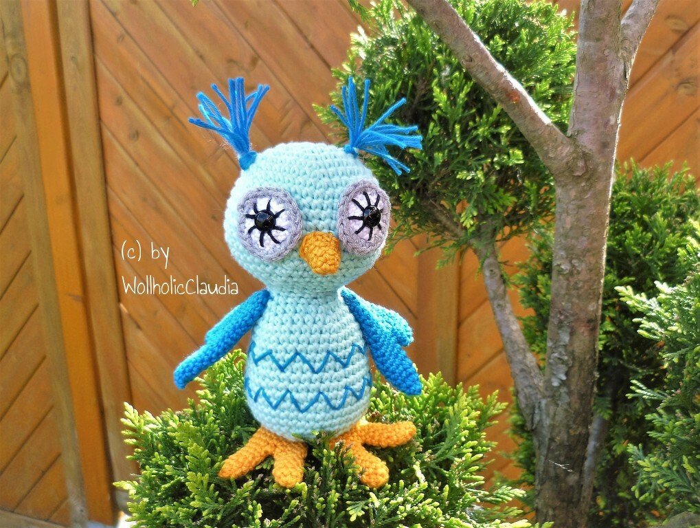 Hellblaue gehäkelte Amigurumi-Eule mit gelbem Schnabel auf einem grünen Strauch