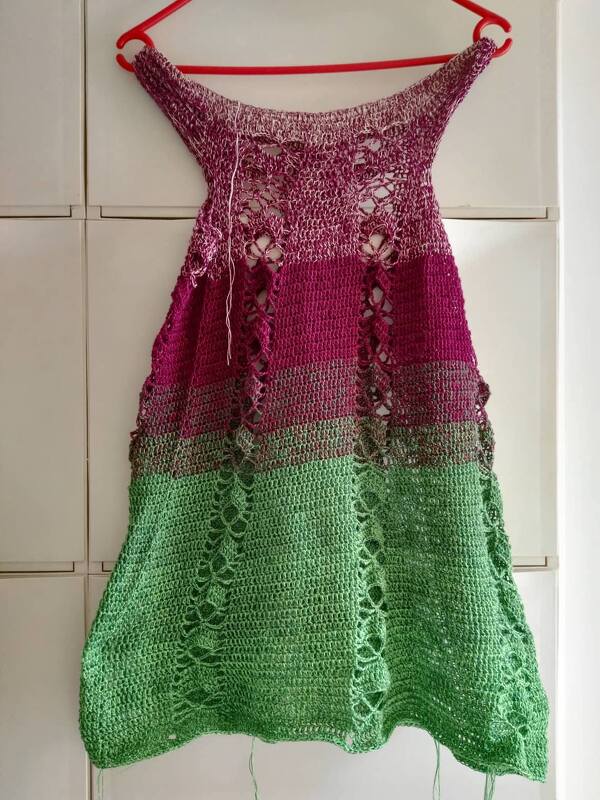 Gehäkelte ärmellose Tunika in abgestuften Pink- und Grüntönen mit vertikalen Lochmustern, an einem roten Kleiderbügel hängend