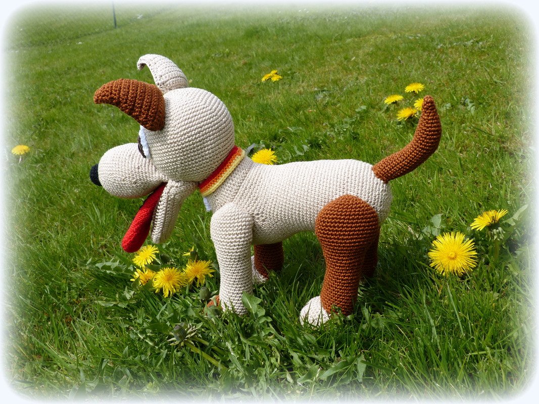 Gehäkelter Amigurumi-Hund in Beige mit braunen Ohren, Pfoten und Schwanz und gestreiftem Halsband auf grüner Wiese mit Löwenzahn.