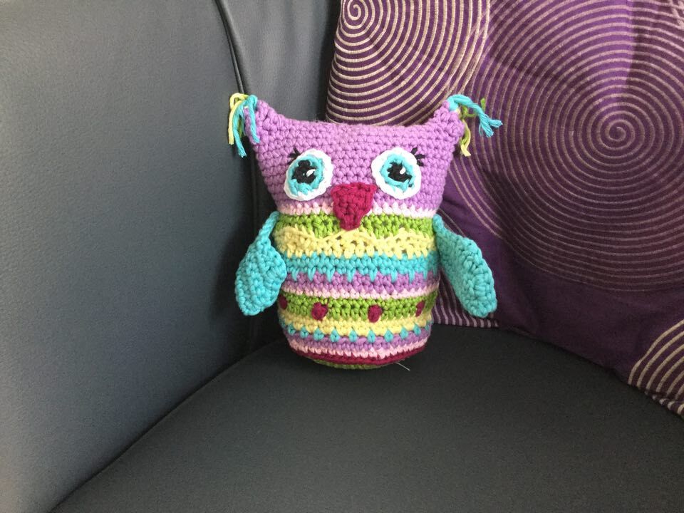 Farbig gehäkelte Eule mit violettem Kopf, gestreiftem Körper und türkisfarbenen Flügeln auf einem Sofa neben einem gemusterten Kissen