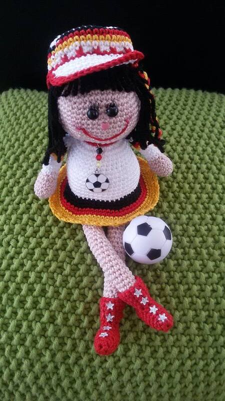 Gehäkelte Amigurumi-Puppe in Schwarz-Rot-Gold mit Mütze, Rock und Fußball, sitzend auf grünem Hintergrund
