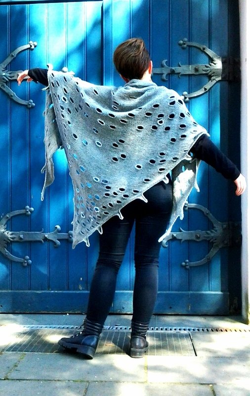 Person von hinten trägt ein graues, grob gestricktes Dreieckstuch mit vielen Lochöffnungen vor einer blauen Tür.