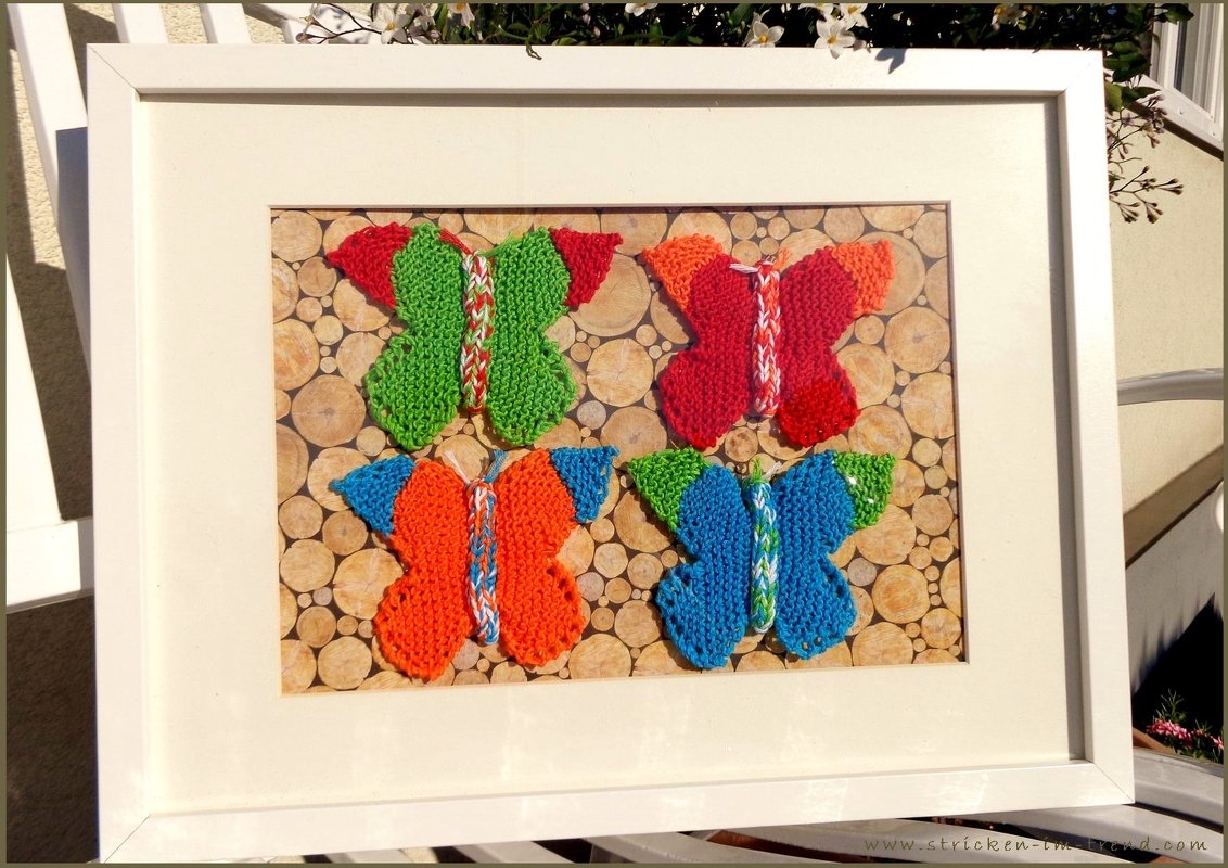 Weißer Bilderrahmen mit vier gestrickten Schmetterlingen in Grün, Rot, Orange und Blau auf Holzscheiben-Hintergrund