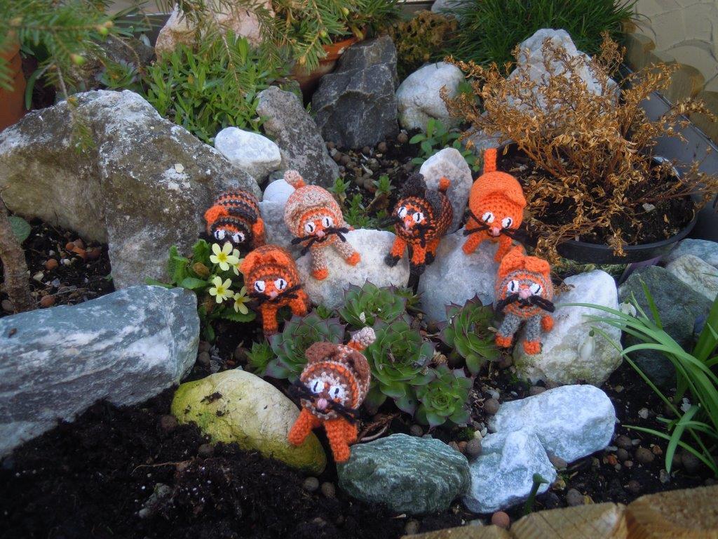 Mehrere kleine gehäkelte orange-braune Katzen-Amigurumi mit Schnurrhaaren und Schwänzen auf Steinen