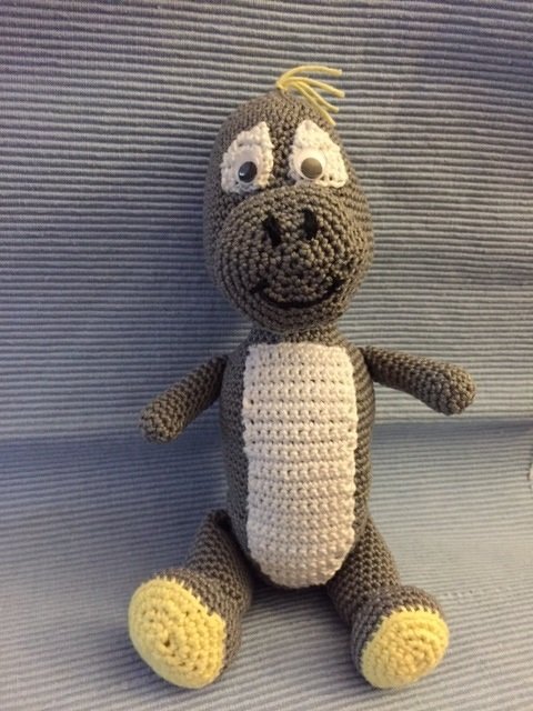 Sitzender, gehäkelter grauer Dinosaurier mit weißem Bauch und gelben Akzenten vor gestreiftem Stoff