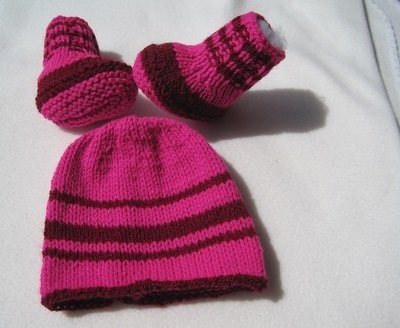 Gestrickte Babymütze und zwei Babyschühchen in Pink mit dunkelroten Streifen auf hellem Hintergrund.