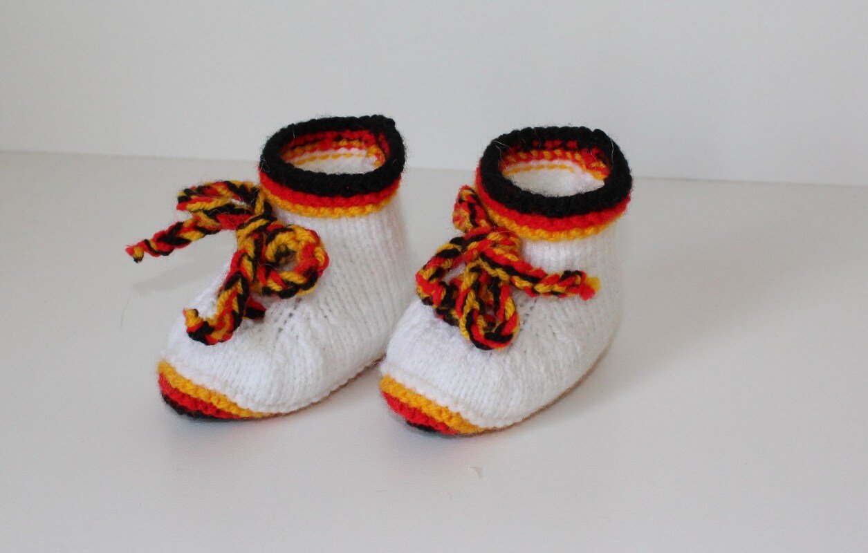 Weiß gestrickte Babyschuhe mit schwarzem, rotem und gelbem Rand und geflochtenen Schnürbändern