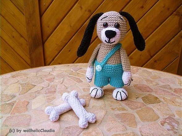 Gehäkelter beiger Amigurumi-Hund mit schwarzen Ohren und türkiser Latzhose auf einem Mosaiktisch; zwei weiße Häkelknochen davor, Holzwand im Hintergrund.