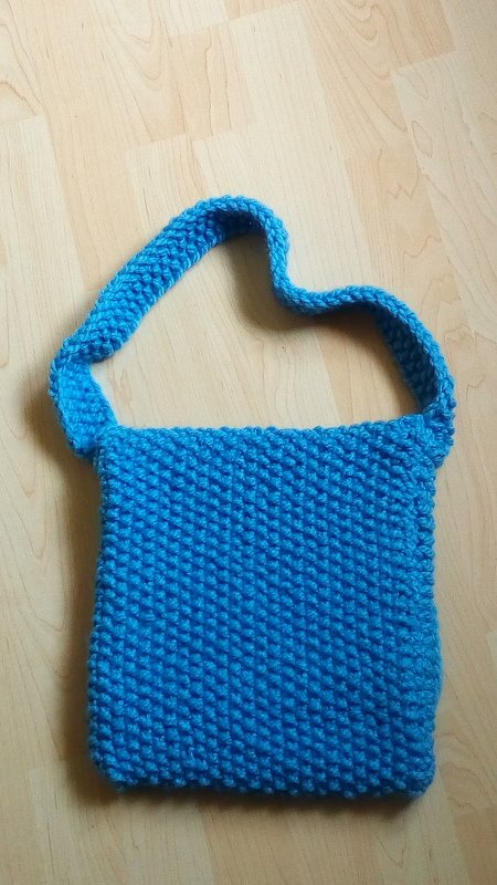 Handgestrickte blaue Umhängetasche mit breitem Schulterriemen auf hellem Holzuntergrund