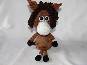 Gehäkeltes Amigurumi-Pferd in Braun mit weißer Schnauze und dunkler Mähne, sitzend vor hellem Hintergrund.