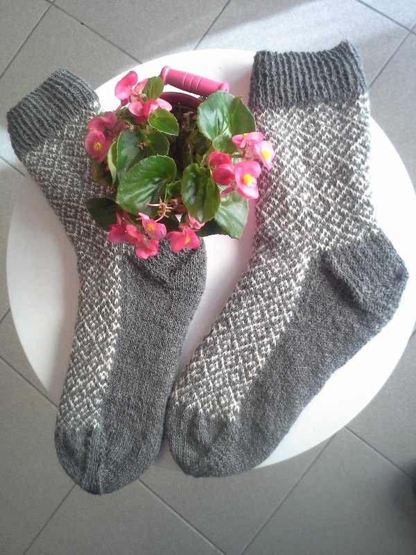 Paar handgestrickter grauer Herrensocken mit weißem Rautenmuster auf rundem weißen Tisch neben Topf mit rosa Blüten