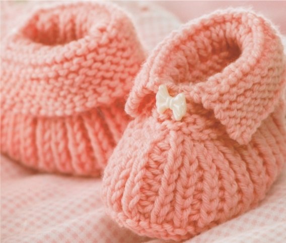 Zartrosa handgestrickte Babyschuhe auf kariertem Stoff mit kleiner weißer Schleife.
