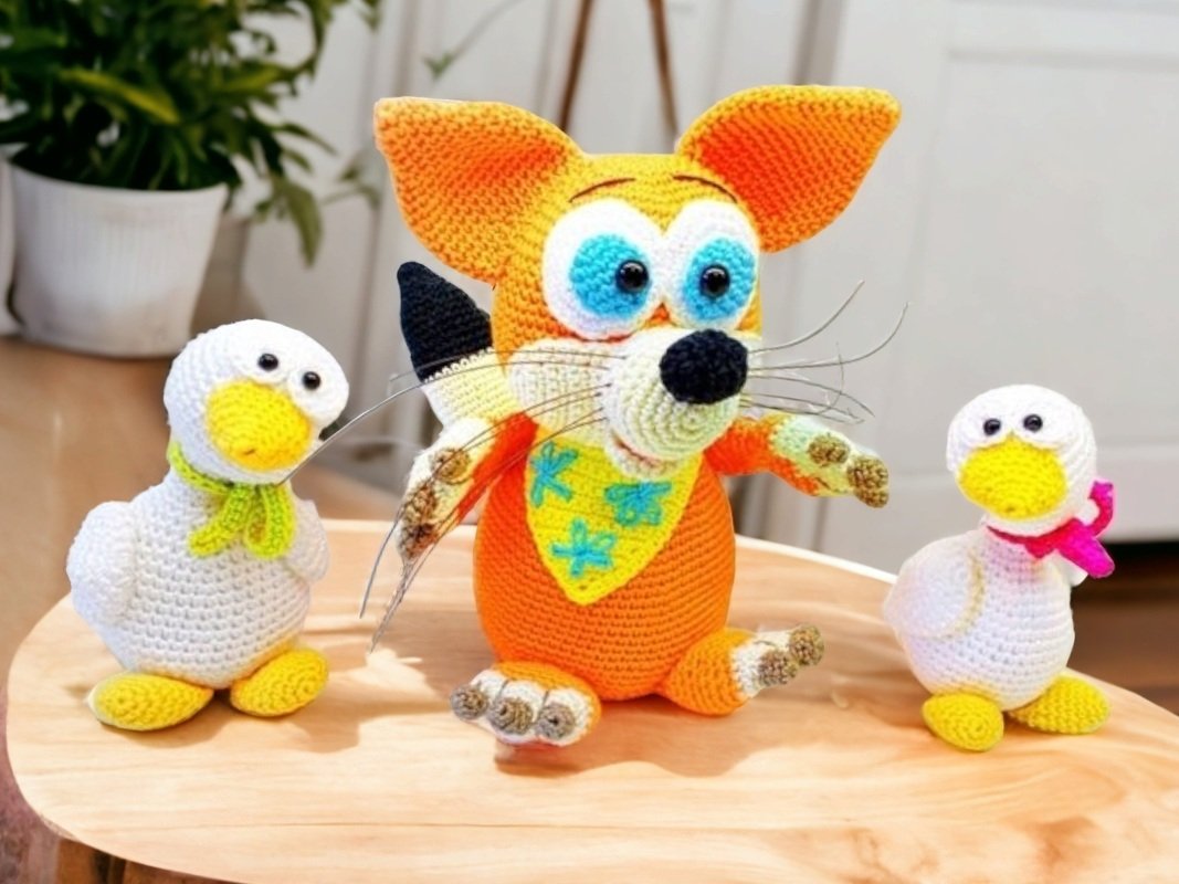 Orange crocheted amigurumi fox with blue eyes and whiskers, flanked by two small white crocheted ducks wearing green and pink scarves.