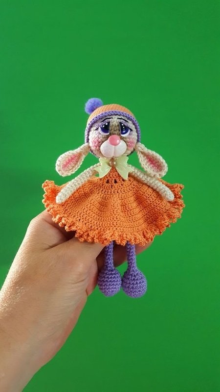 Hand hält einen gehäkelten Amigurumi-Hasen im orangefarbenen Kleid mit lila Bommelmütze vor grünem Hintergrund.