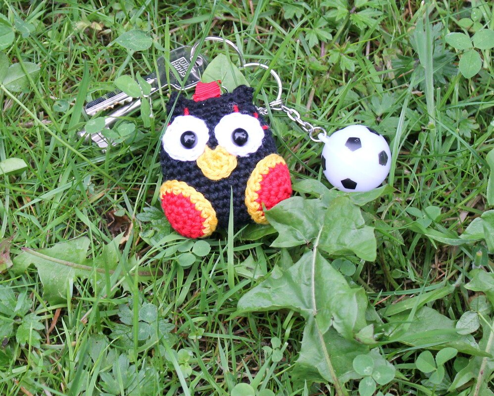 Gehäkelte Eule (schwarz, rot, gelb) als Schlüsselanhänger mit kleinem Fußball-Anhänger auf Gras neben Schlüsseln.