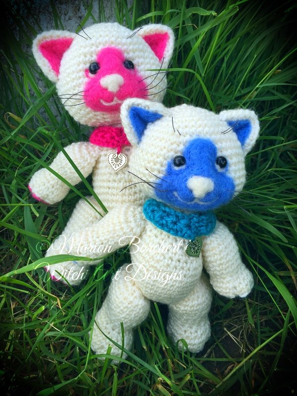 Zwei gehäkelte Amigurumi-Katzen in Creme mit blauem bzw. rosa Gesicht und Halsbändern im Gras