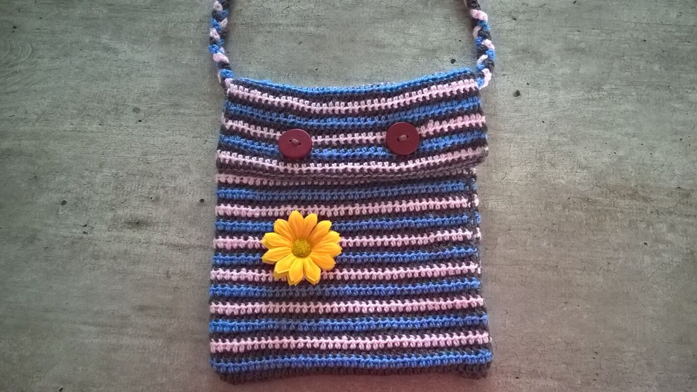 Gehäkelte Henkeltasche mit blau-rosa-grauen Streifen, Klappe, zwei roten Knöpfen und gelber Blume auf grauem, strukturiertem Untergrund.