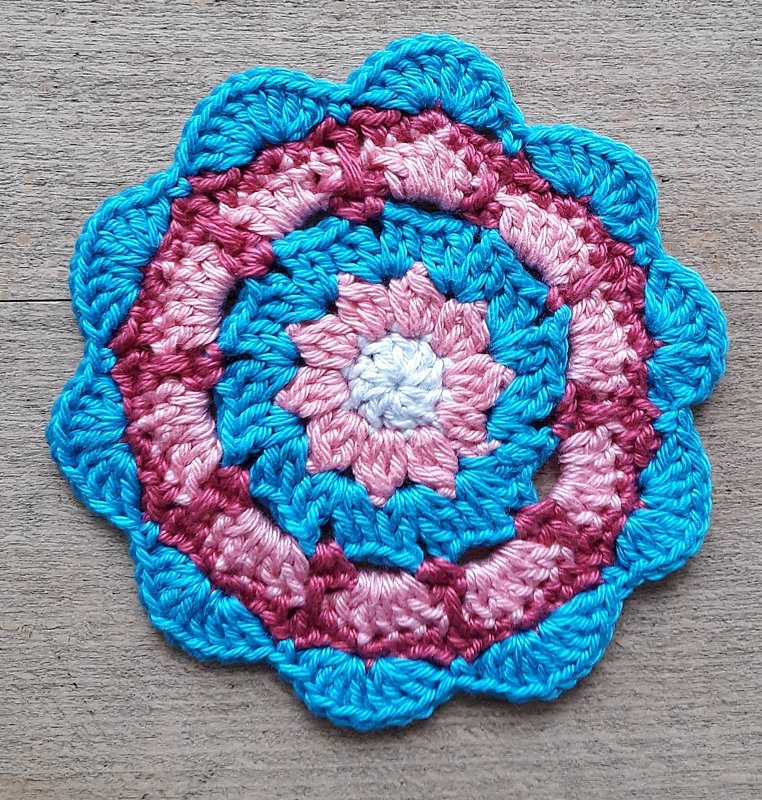 Runder gehäkelter Untersetzer in blau und rosa mit muschelförmigem Rand auf Holz