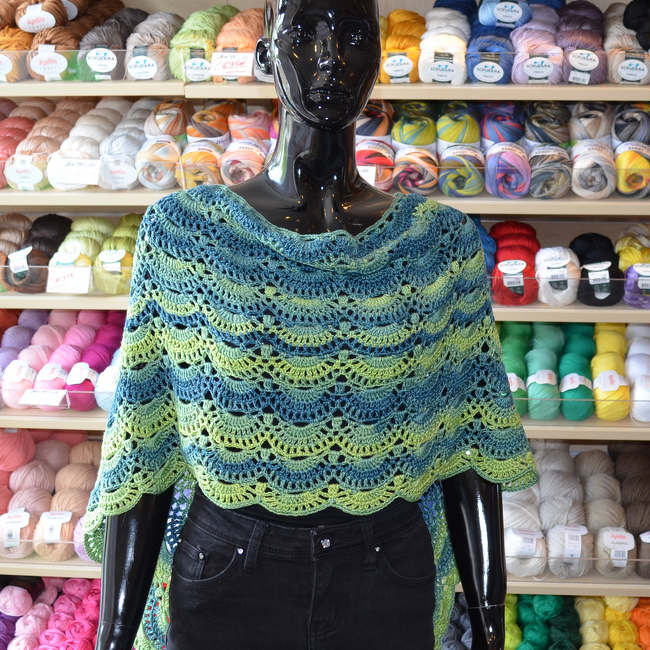 Kurzponcho mit Muschelreihen und gezacktem Saum in Blau‑Grün auf Schaufensterpuppe vor Garnregal