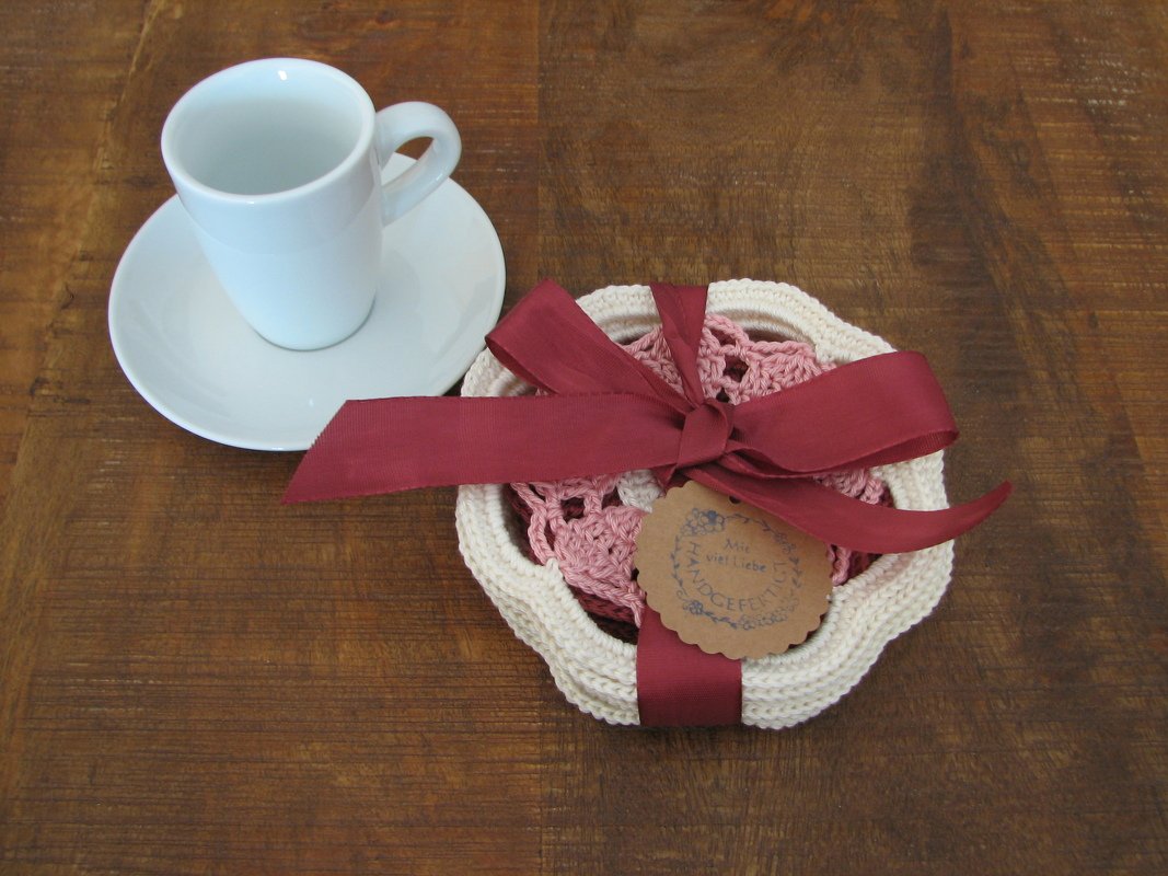 Stapel gehäkelter Untersetzer in Rosa und Creme mit dunkelroter Schleife neben einer weißen Tasse auf einem Holztisch.