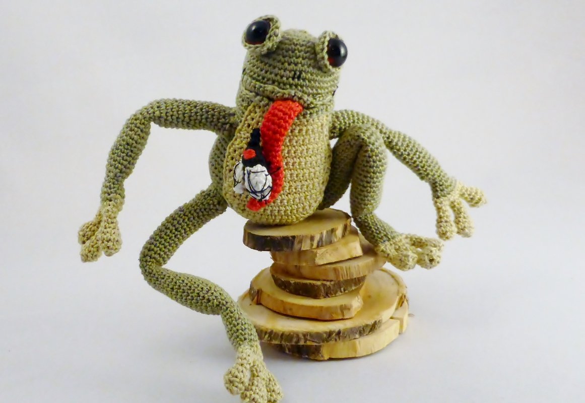 Hellgrüner gehäkelter Amigurumi-Frosch mit roter Zunge und Fliege auf Holzscheiben vor weißem Hintergrund