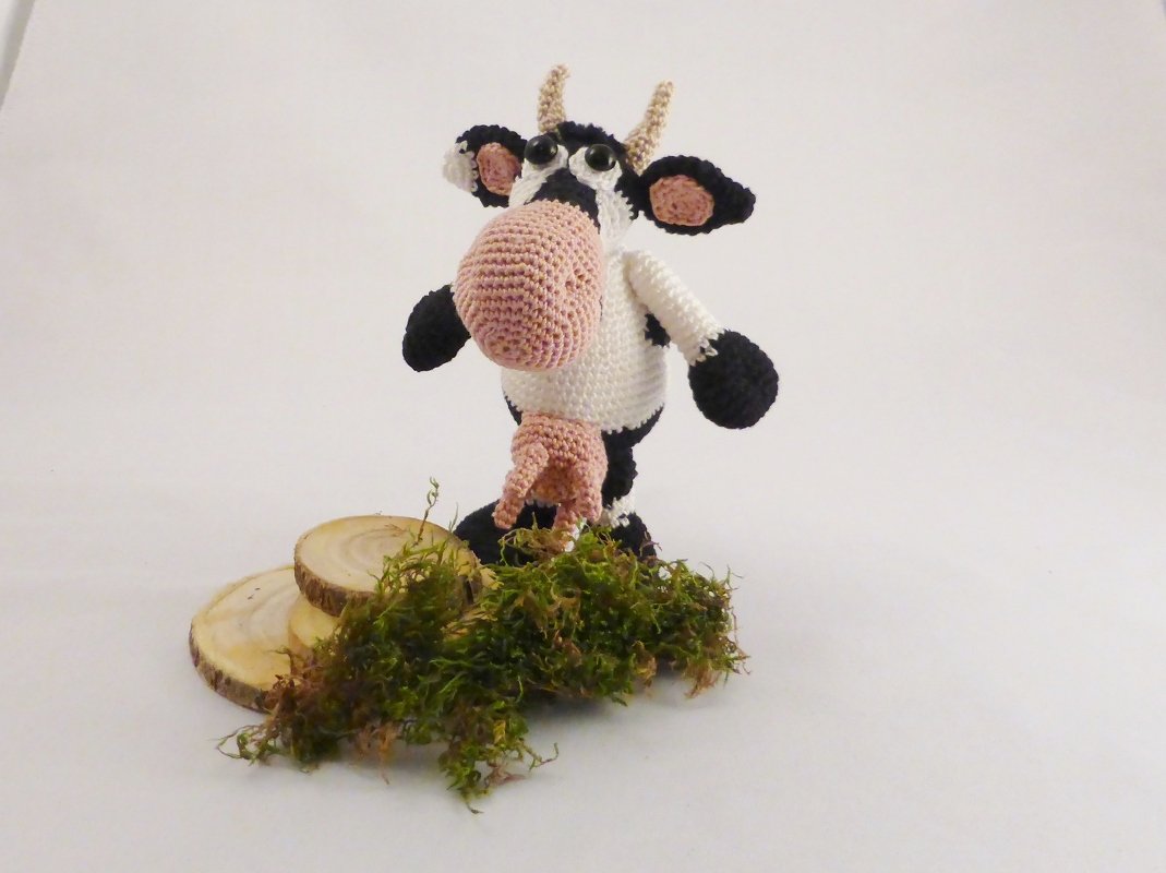 Gehäkelte schwarz-weiße Kuh mit rosa Schnauze und Euter auf Moos und Holzscheiben