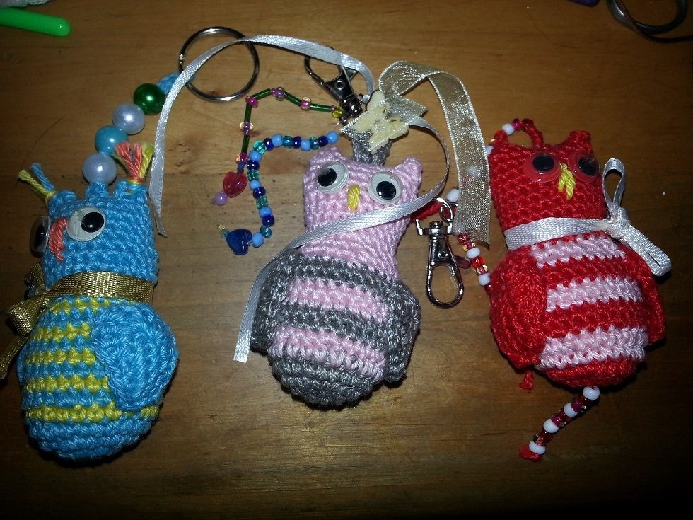 Drei gehäkelte Amigurumi‑Eulen‑Schlüsselanhänger in Blau, Rosa und Rot auf Holzuntergrund