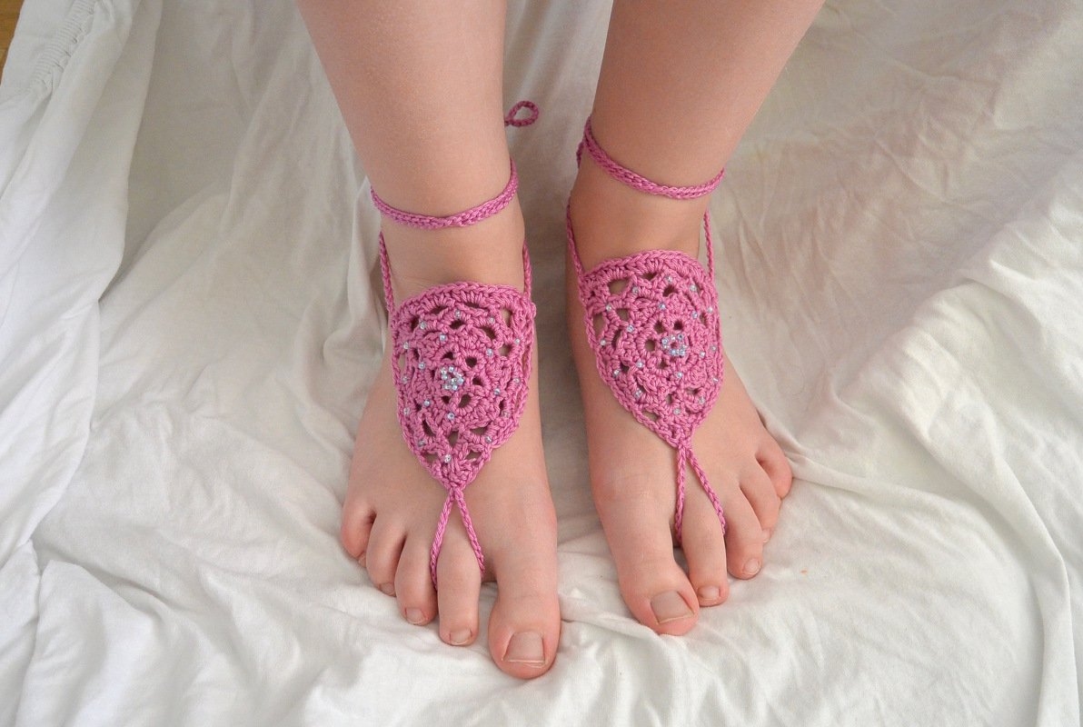 Nahaufnahme von Füßen mit rosa gehäkelten Barfußsandalen mit Perlen auf weißer Decke.