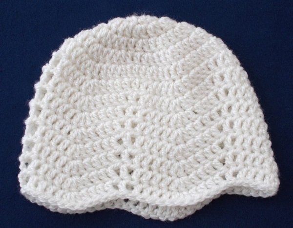 Weiße gehäkelte Babymütze mit Muschelmuster auf dunkelblauem Hintergrund.