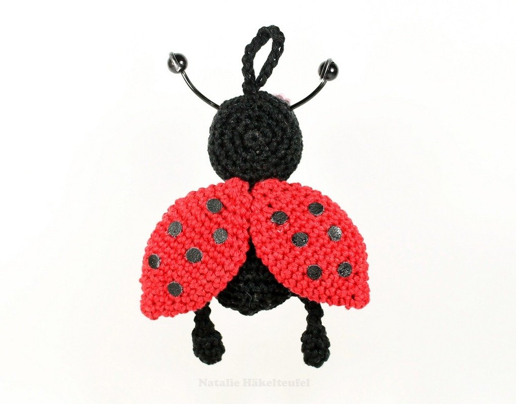 Nahaufnahme eines gehäkelten Marienkäfers in Rot und Schwarz mit Fühlern und Aufhängeschlaufe vor weißem Hintergrund.