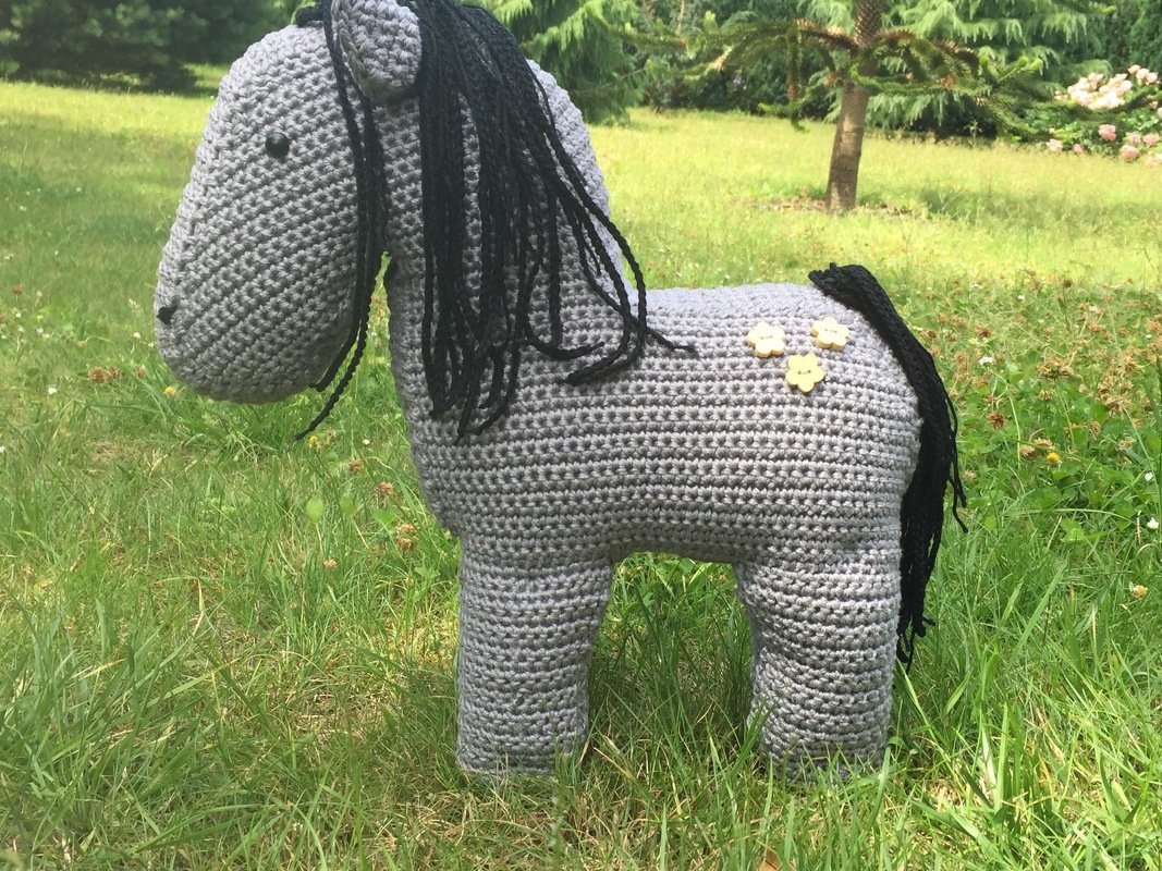 Gehäkeltes graues Pferd mit schwarzer Mähne und drei Blumenknöpfen auf Gras.