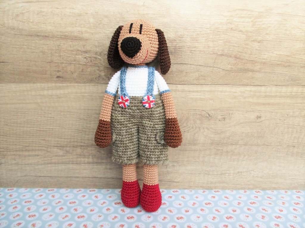 Gehäkelter Amigurumi-Hund mit Latzhose, blauen Trägern und roten Schuhen vor Holzuntergrund
