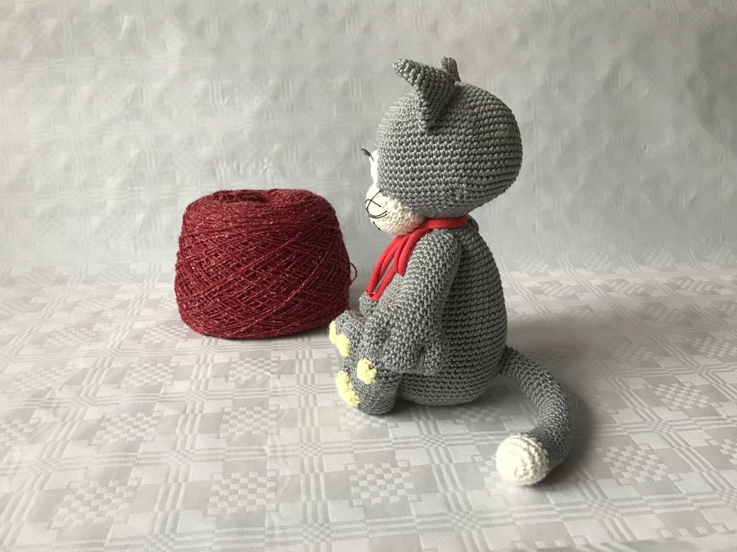 Gehäkelte graue Amigurumi-Katze mit rotem Schal neben roter Garnrolle, seitliche Ansicht