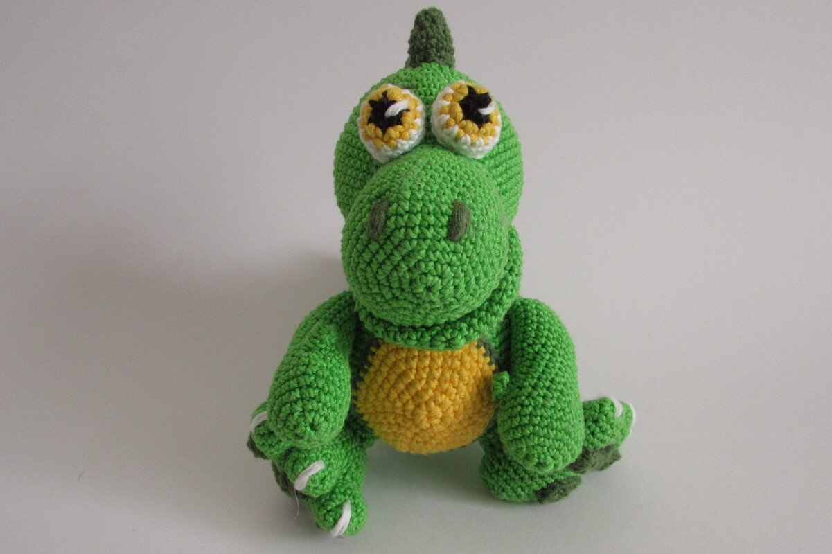 Grüner gehäkelter Amigurumi-Dinosaurier mit gelbem Bauch und großen Augen vor hellem Hintergrund.